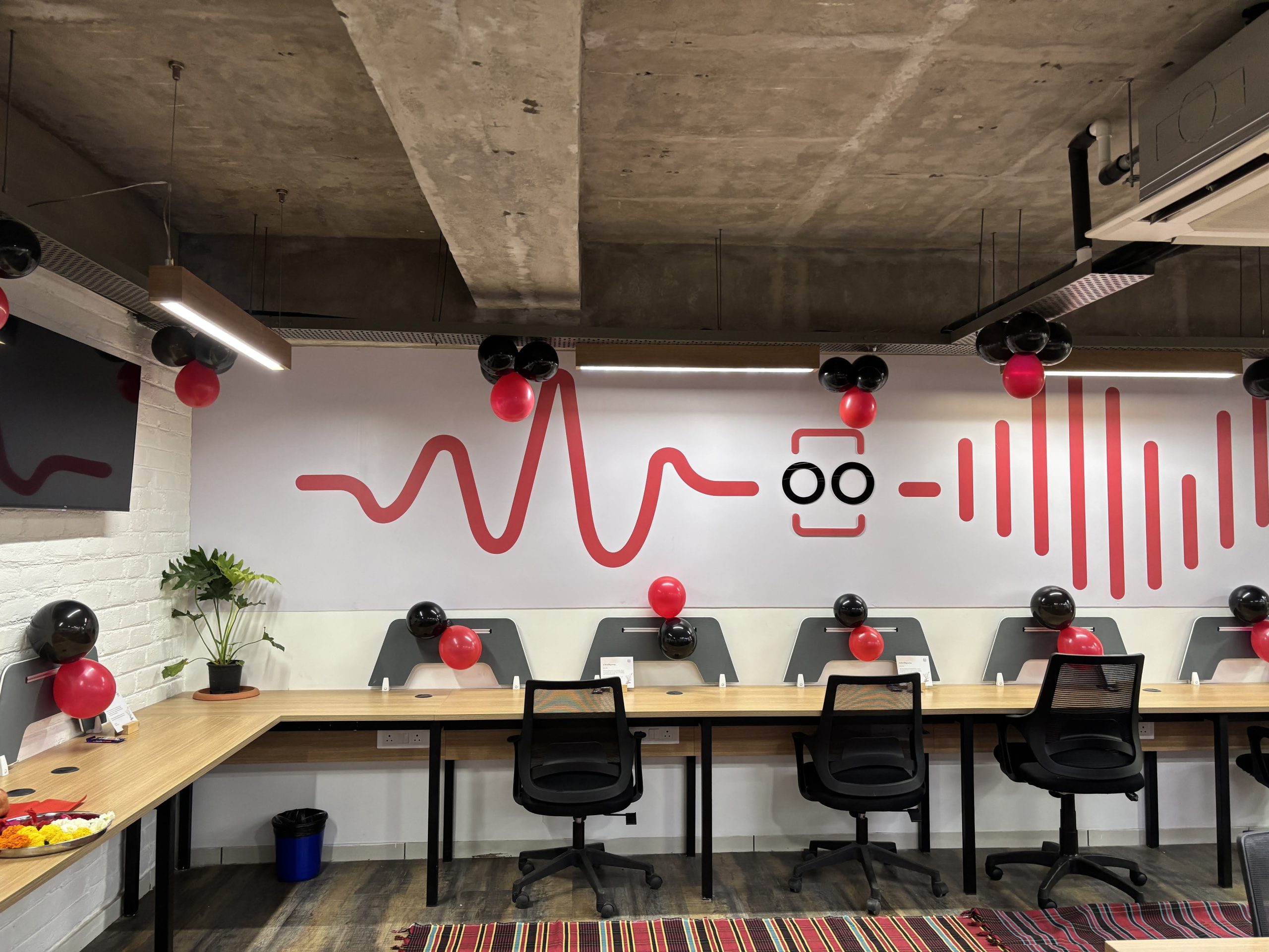 Open office area with row of desks and black chairs, red and black balloons along the wall, and a white wall with red graphic patterns under a concrete ceiling.