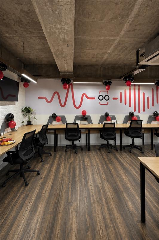 Open-plan conference room with long wooden desks, black chairs, red and black balloons, and a mural of red abstract waves with a playful face graphic on the wall.
