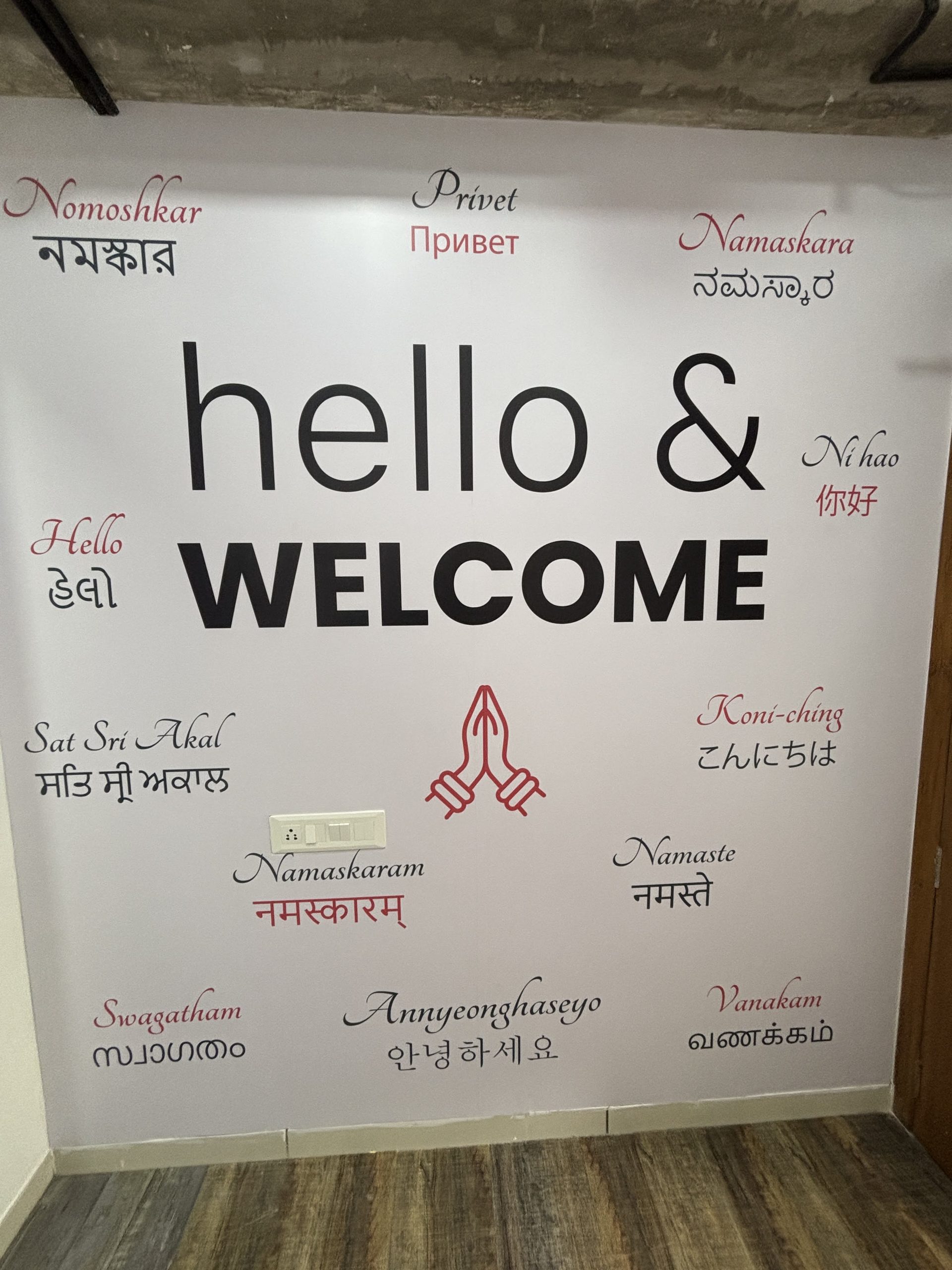 Wall graphic with the words 'hello' and 'WELCOME' in large black type, surrounded by 'hello' in many languages and a red praying-hands symbol at center bottom. Floor and exposed ceiling visible.