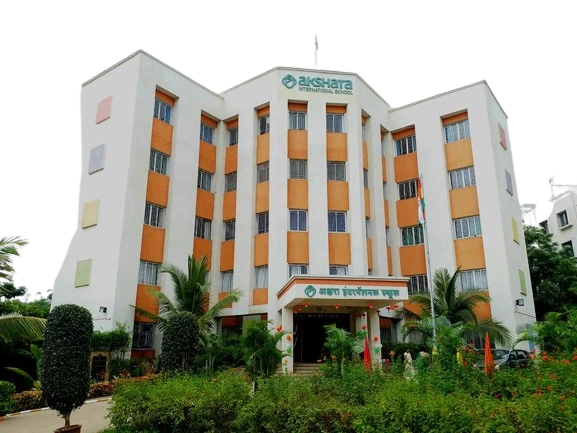Exterior view of Akshara International School building, a white multi-story structure with orange vertical panels, surrounded by palm trees and a landscaped entrance.
