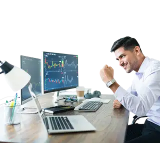 Businessperson smiling and fist-pumping at a desk with two monitors showing stock charts and a laptop nearby